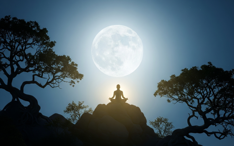 Silhouette of a person meditating on a mountaintop beneath a radiant full moon framed by trees.
