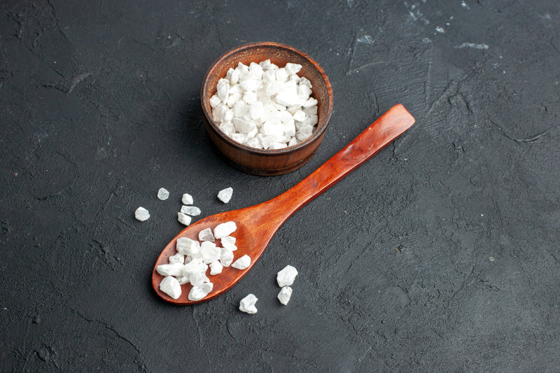 A picture of white Camphor crystals in bowl and spoon placed on the dark surface