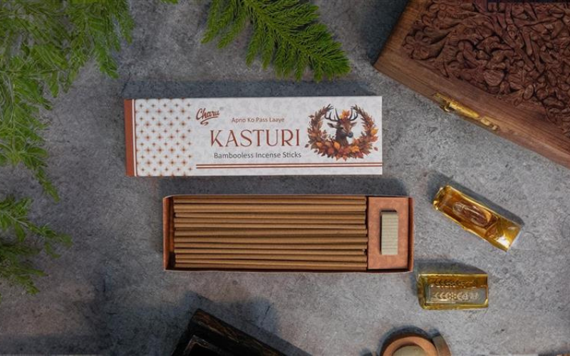 Charu perfumery  bamboo less  kasturi incense sticks with an open box, greenery, carved wood, and aromatic oil bottles on a gray surface