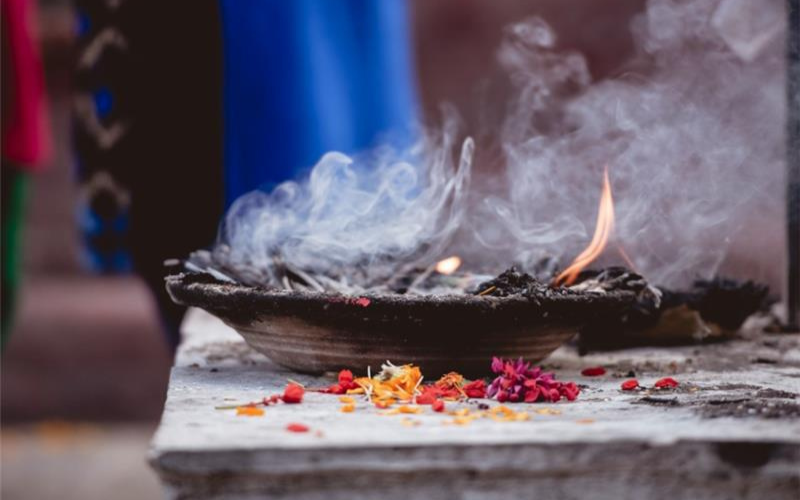 Clay incense burner with smoke and flame, surrounded by scattered flower petals on a stone surface.