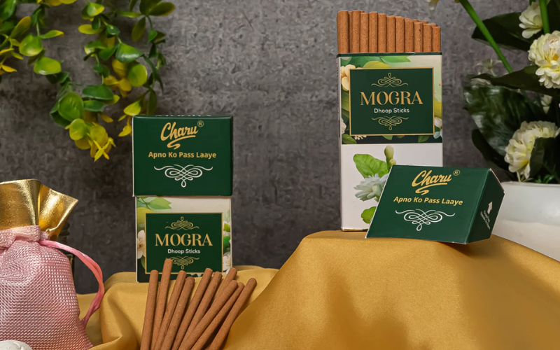 Pack of 27 Charu Mogra Dhoop Sticks displayed with boxes, decorative flowers, and wellness items on a yellow cloth backdrop.